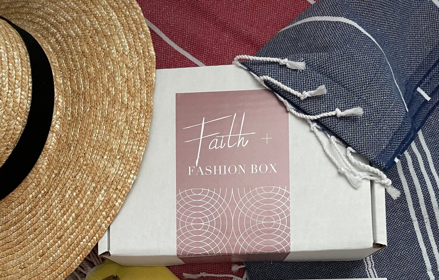 A white box labeled "Faith + Fashion Box" is placed on a textured fabric, next to a straw hat and a folded cloth with tassels.