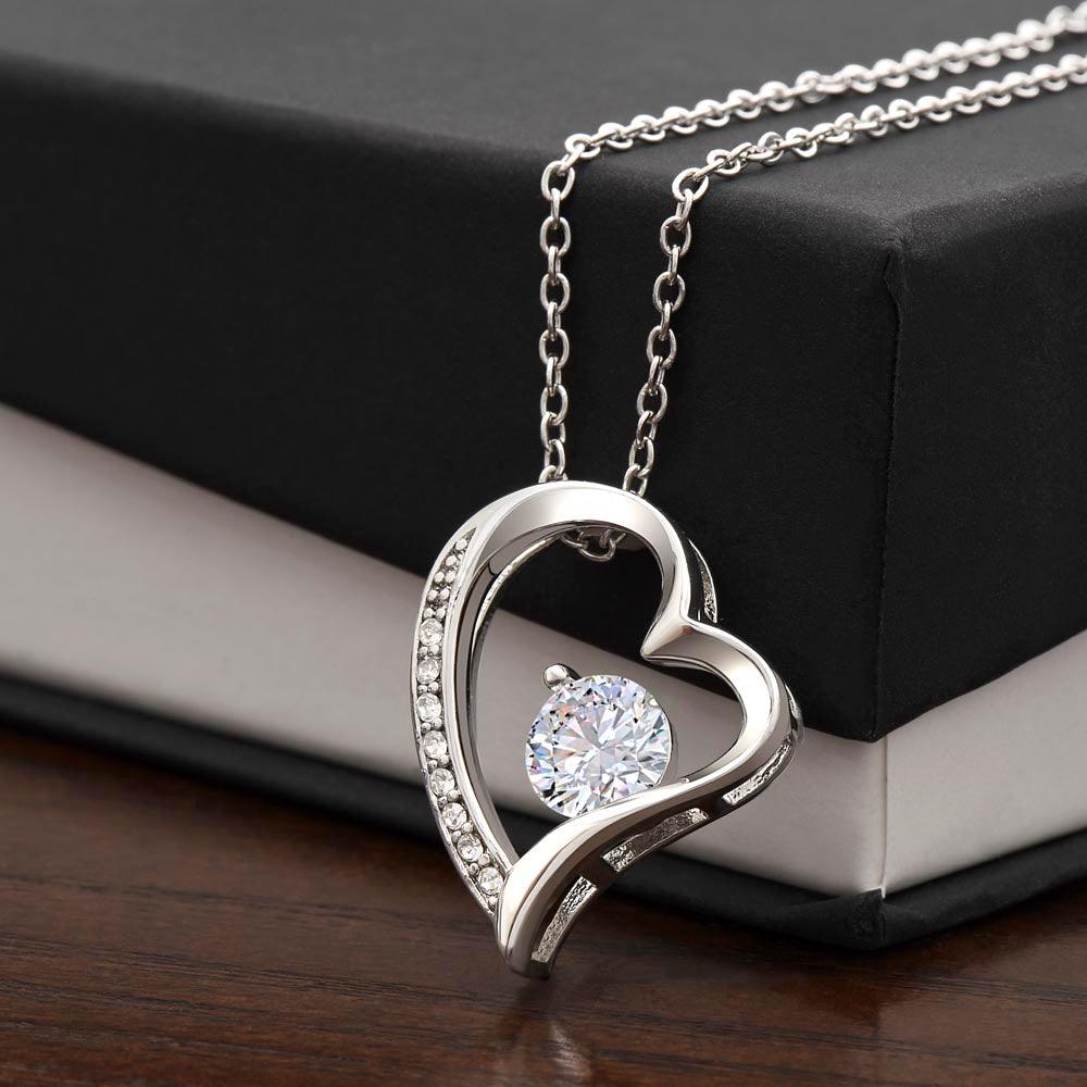 The Forever Love Necklace by ShineOn Fulfillment showcases a silver heart-shaped design with a central diamond pendant and a white gold finish, elegantly presented in a black and white box on a wooden surface.