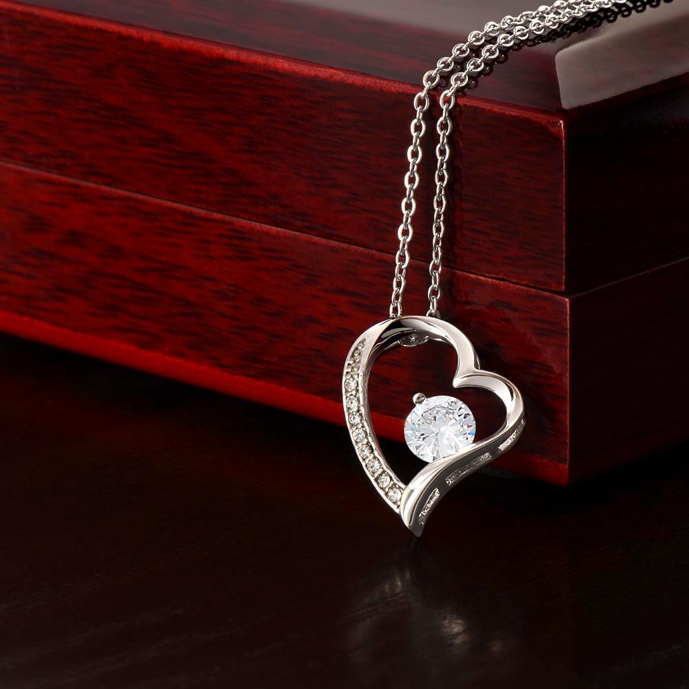 The Forever Love Necklace by ShineOn Fulfillment features a silver heart pendant adorned with a CZ crystal, elegantly displayed on a polished wooden surface. Its white gold finish enhances its timeless beauty and charm.